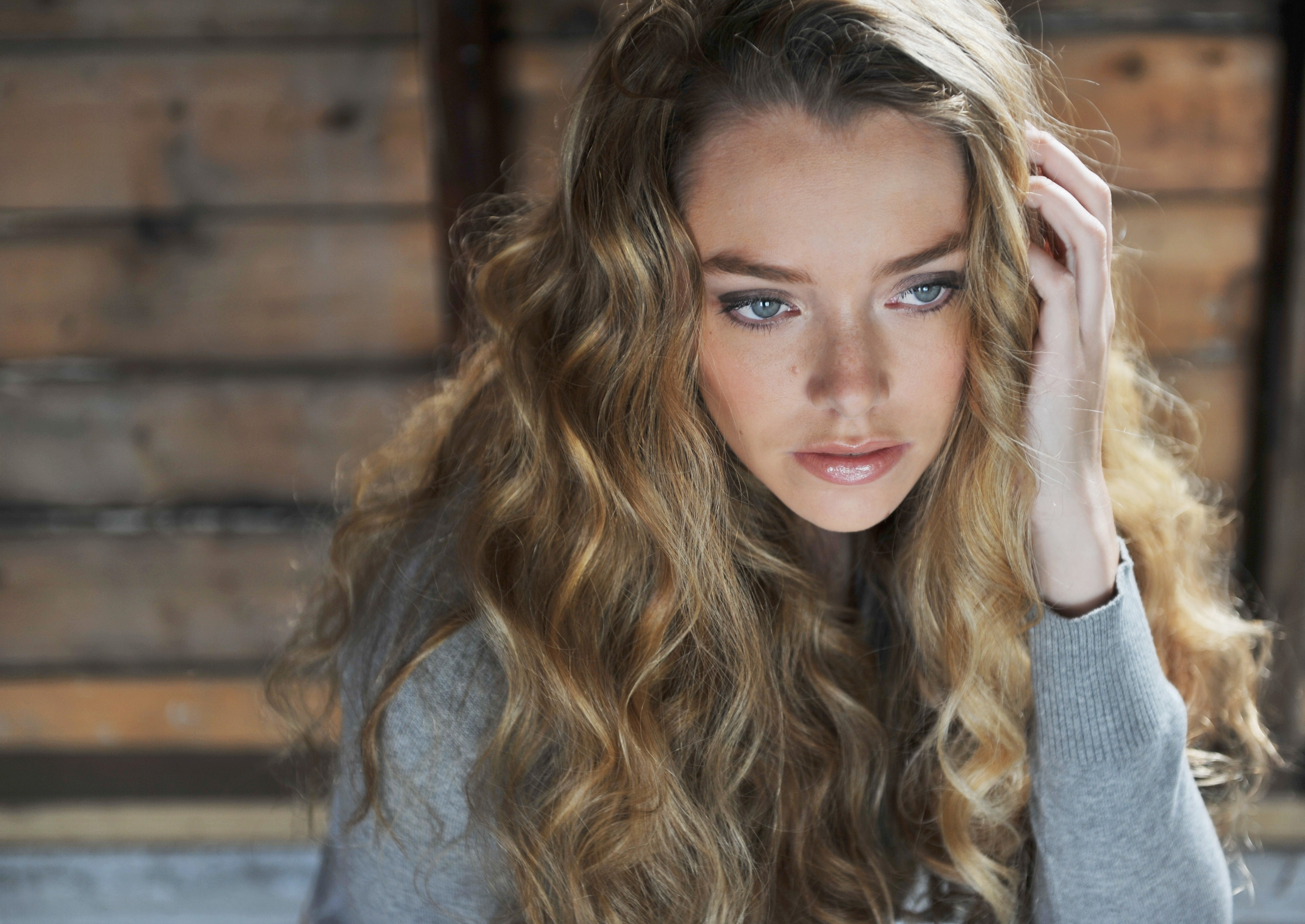 Soft-light portrait of Caitlin against a wood-plank wall — long, loose beach waves framing her face, natural makeup with smoky eyes. Hair by Angeleah Daidone.