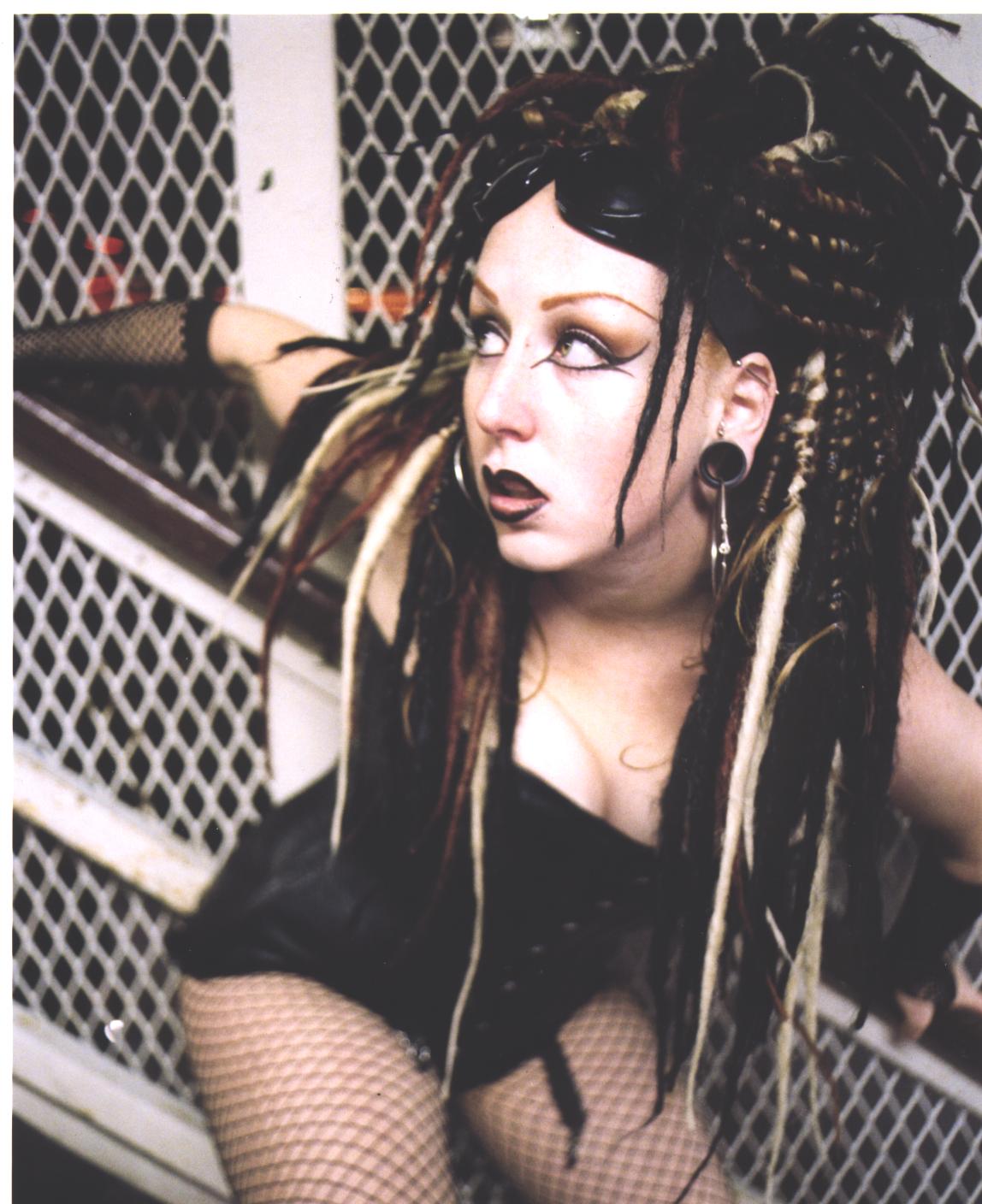 Tamale Sepp in dramatic gothic styling — long multi-toned synthetic dreadlocks in black, red and blonde, heavy eye makeup, dark lipstick and large ear gauges, posed against a chain-link backdrop. Hair by Angeleah Daidone.