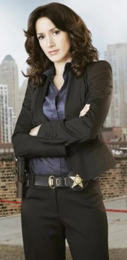 Promotional photo of Jennifer Beals as a detective on The Chicago Code, wearing a black blazer with badge and holster at her belt, the Chicago skyline behind her.