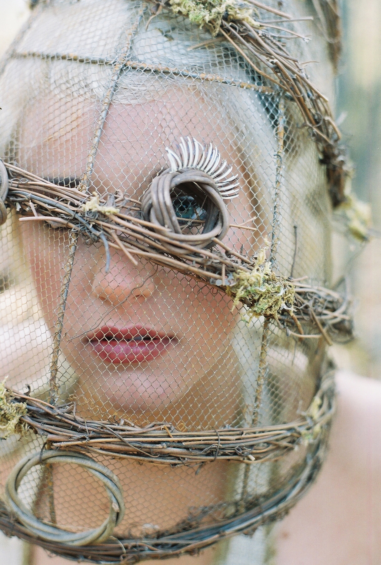 Conceptual portrait of Nicole behind a veil of chicken wire and twigs, with a metallic feathered eye decoration and dark lipstick. Hair by Angeleah Daidone.