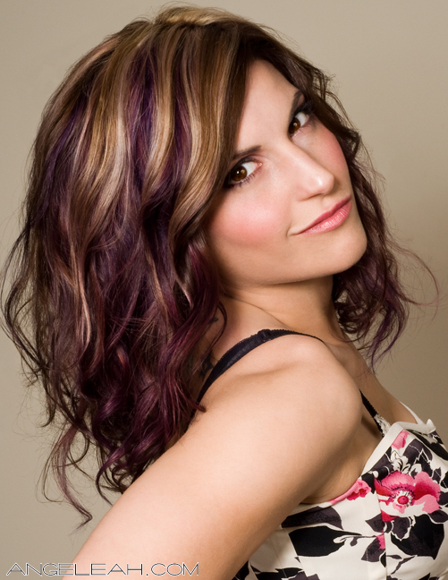 Three-quarter portrait of Racheal — shoulder-length wavy brunette hair with blonde and purple highlights, soft pink makeup, wearing a black-and-white floral top. Hair by Angeleah Daidone.