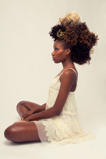 Side-profile portrait of Unique — a dramatic sculpted natural-hair updo with gold accents — seated in a sheer cream lace dress against a soft neutral backdrop. Hair by Angeleah Daidone.